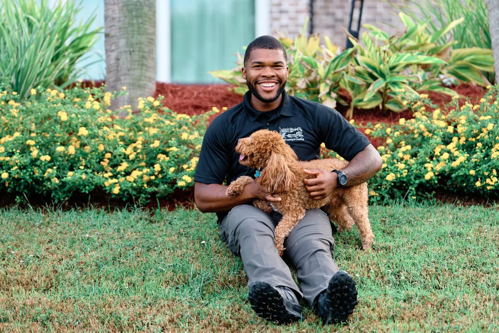 Man sitting on grass holding a small brown dog, smiling.