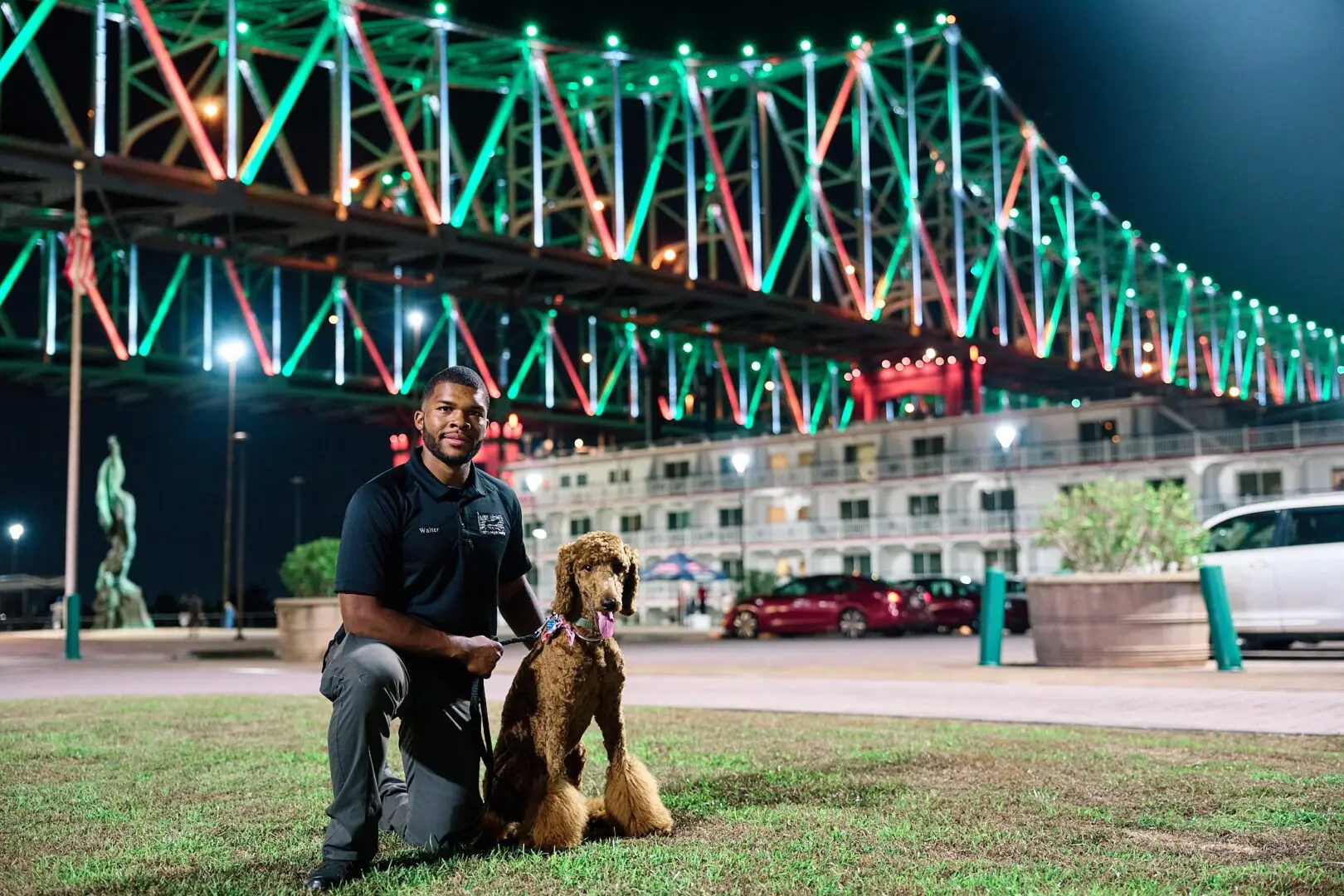 Man kneeling with his dog in front of a brightly lit bridge at night.