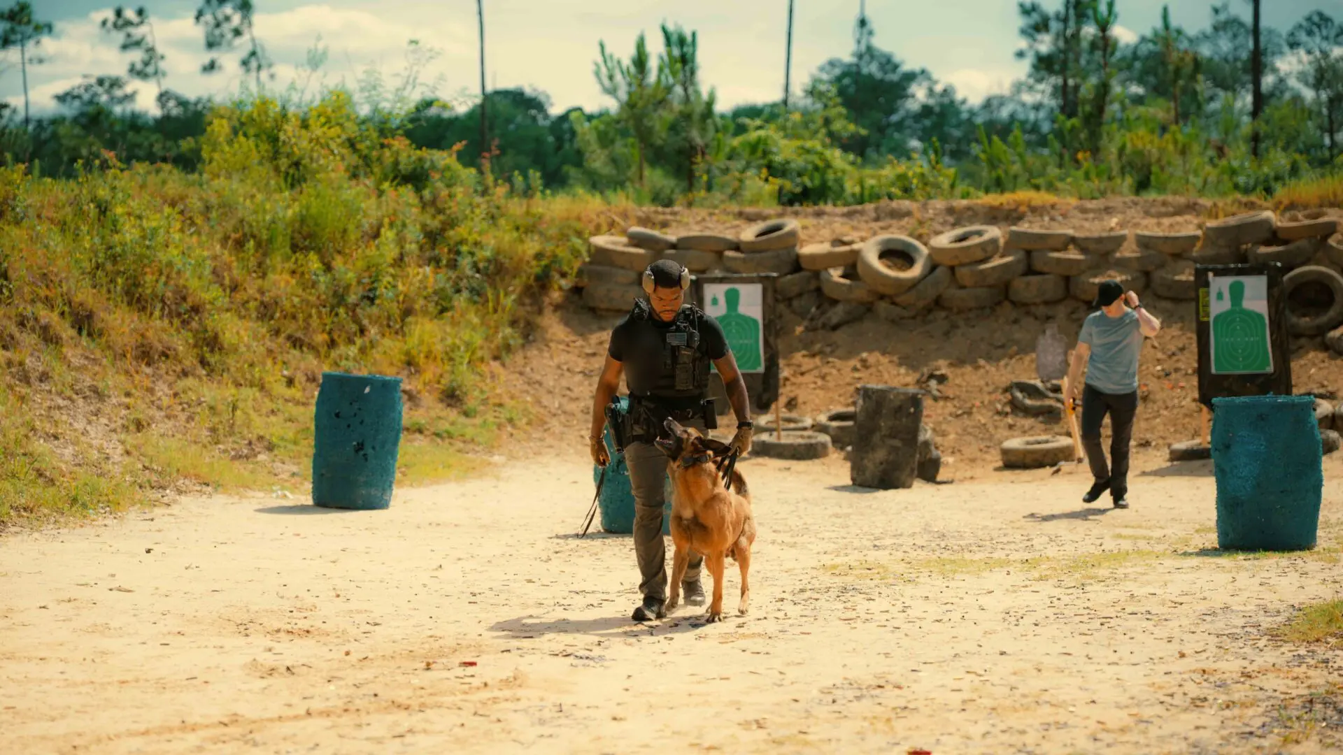 A person with a dog in a rugged outdoor area with tires and barrels.