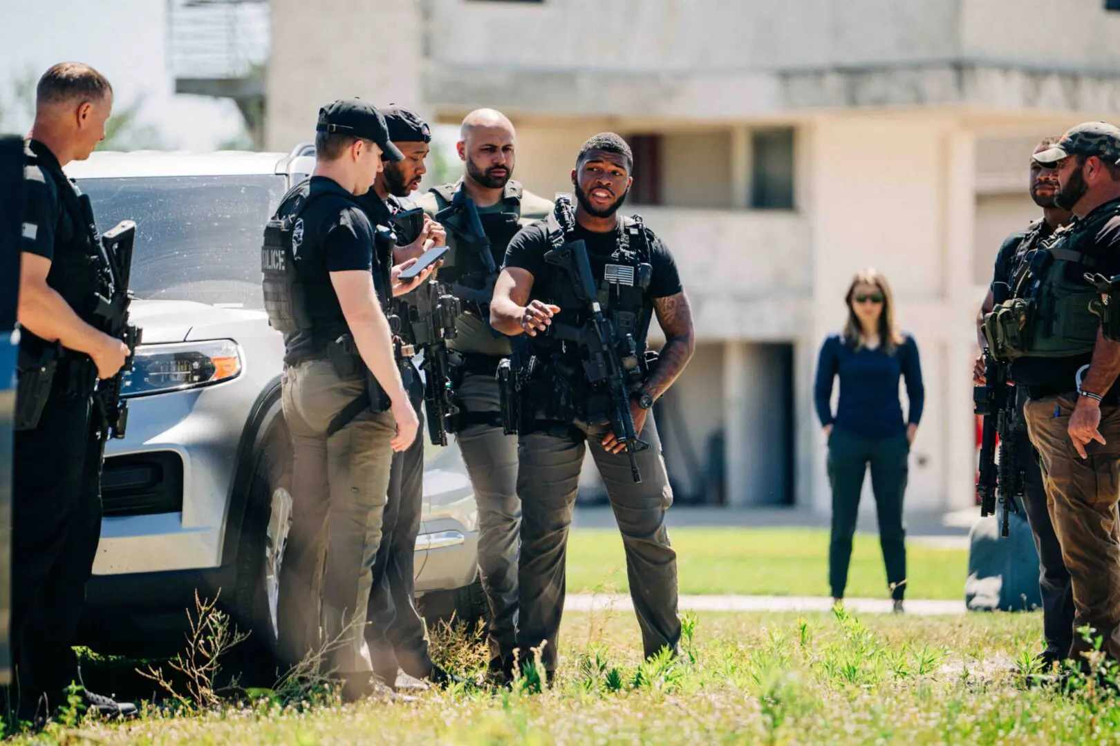 Three police officers talking on a grassy area with a woman in the background.