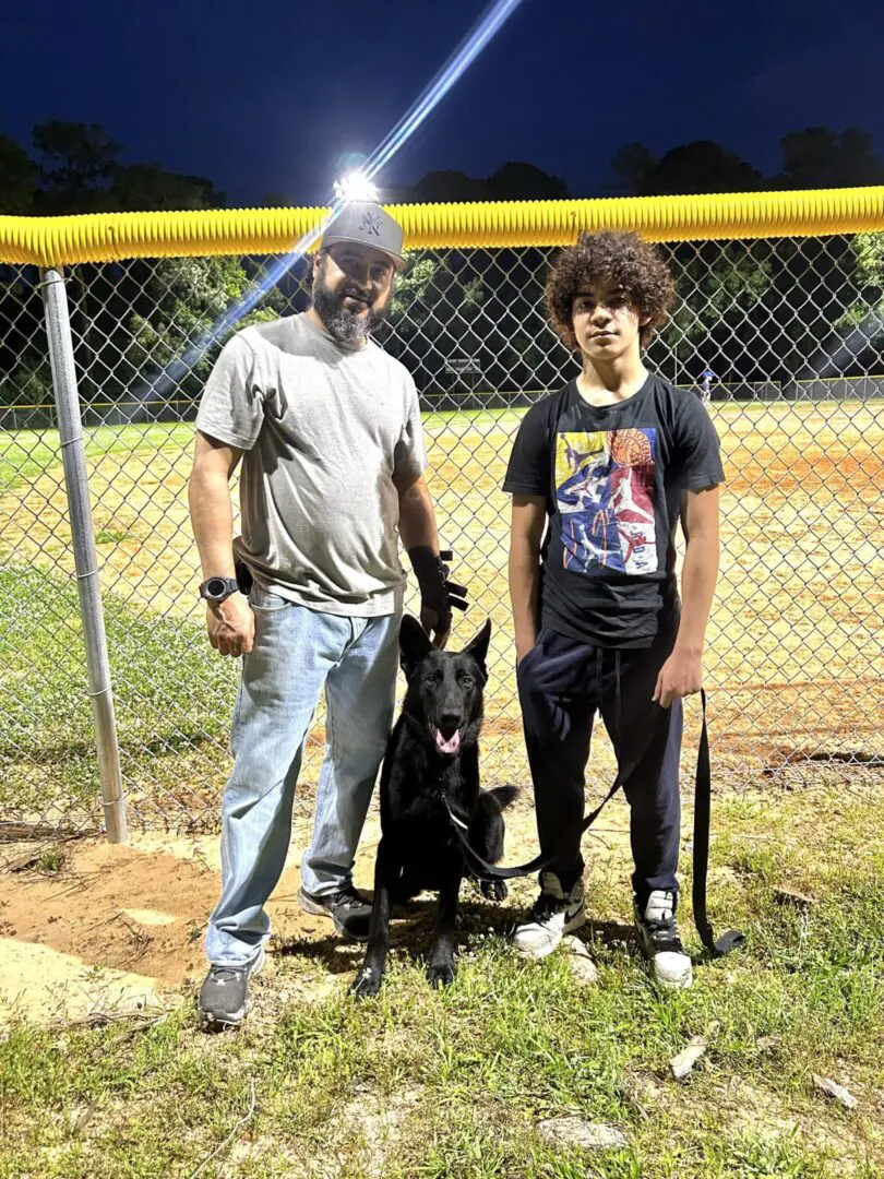 Two young men with a black dog at a baseball field at night.