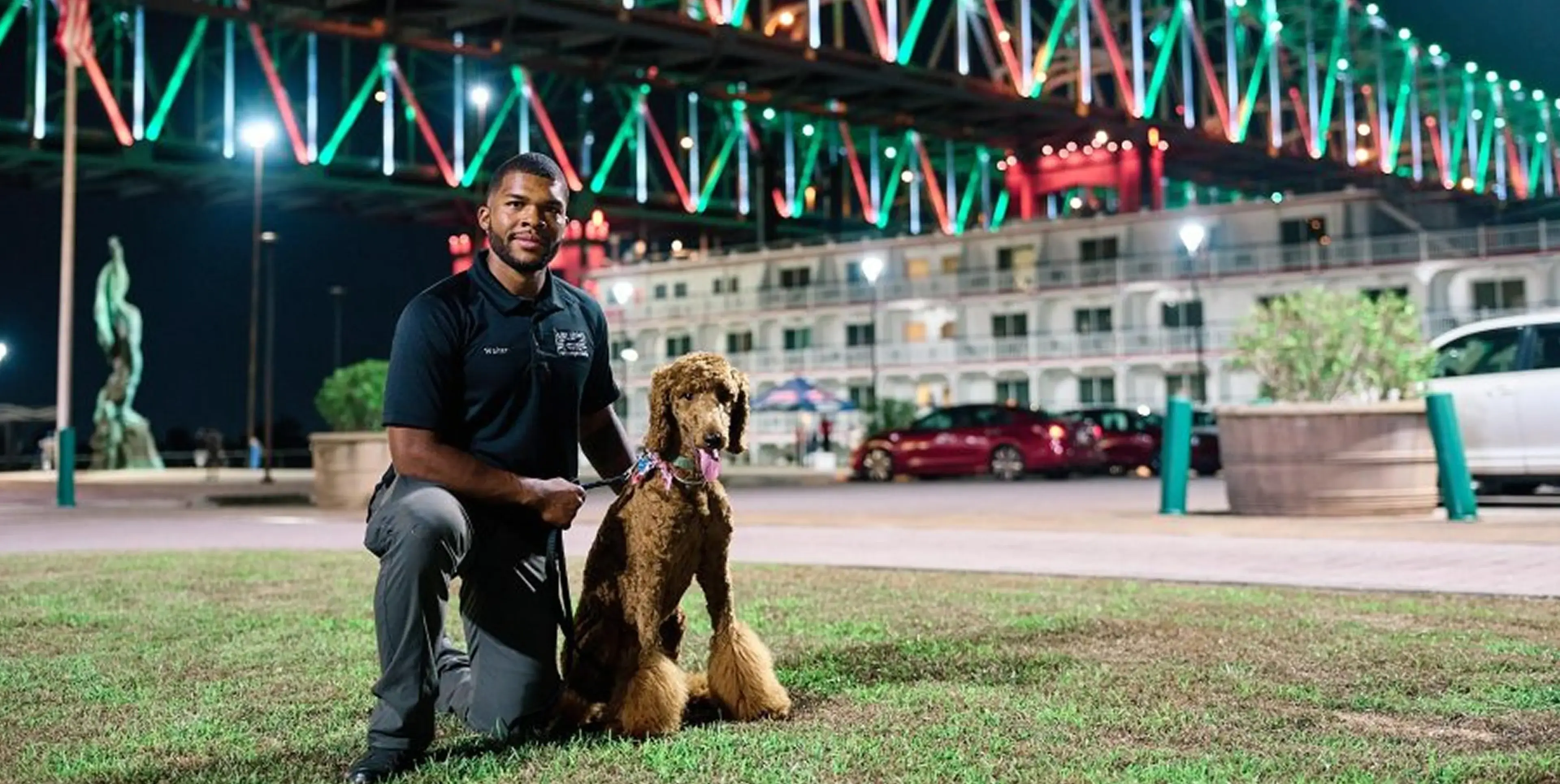 Man posing with a dog in front of a festive, lit-up building at night.