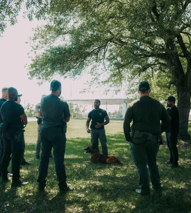 People gathered outdoors under trees, listening to a speaker.