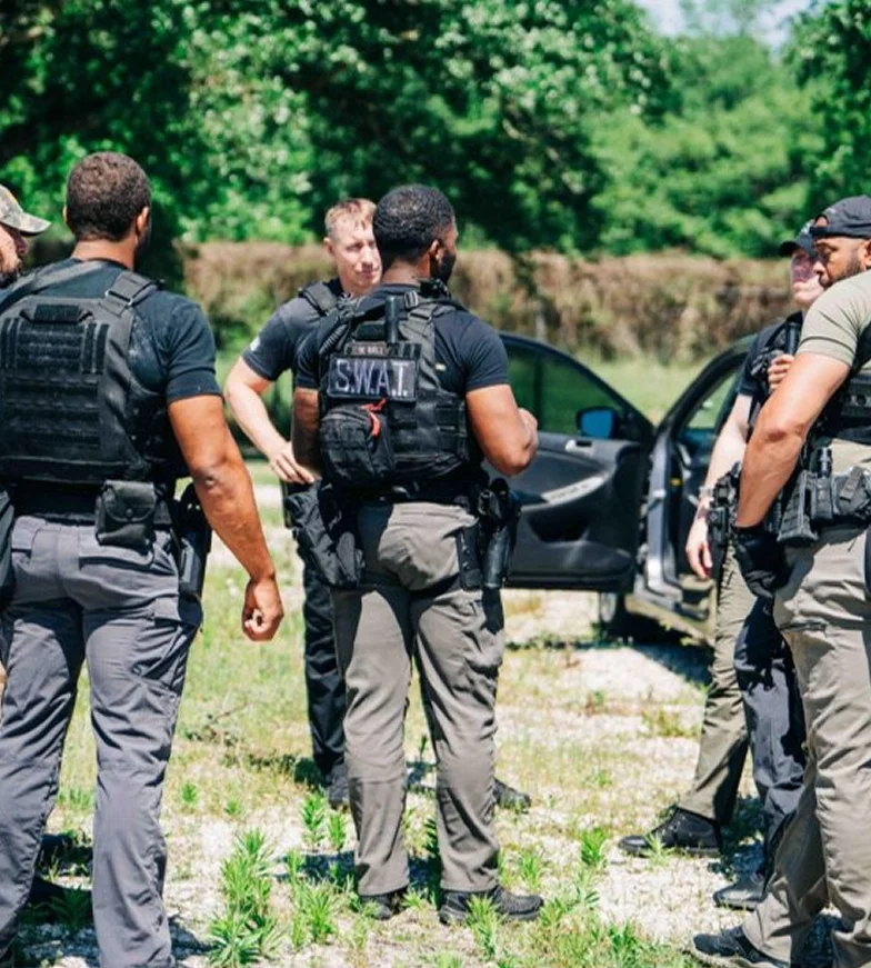 Law enforcement officers gather near a damaged vehicle in a wooded area.